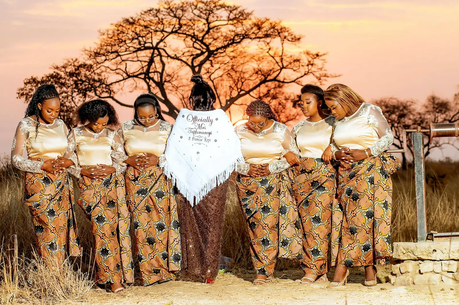A company of women in gold tops and patterned skirts, standing together outdoors.