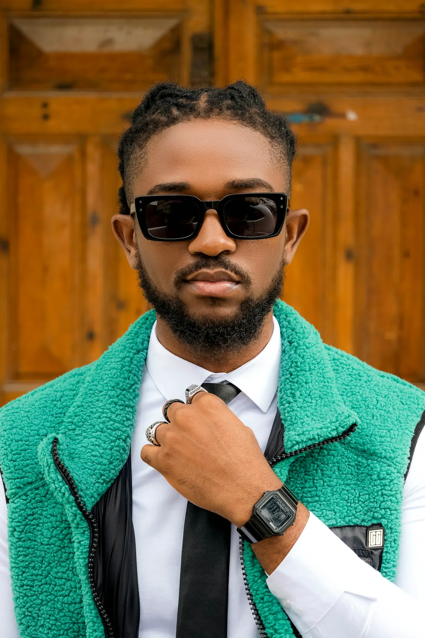 A man in a green tailored jacket, black tie, and sunglasses, hand at the collar.