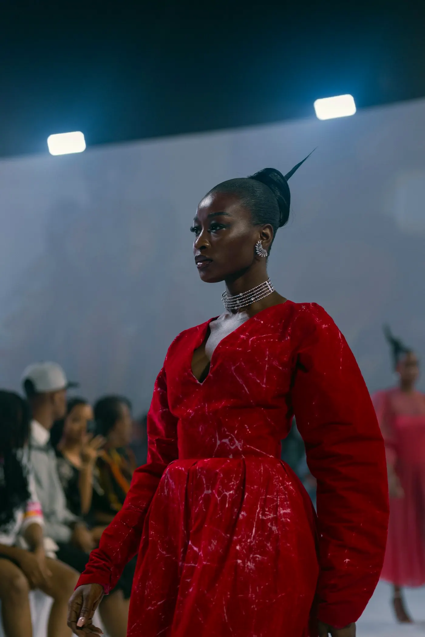 A model in a red gown standing at the end of a runway, audience on either side.